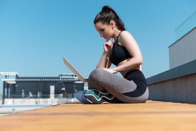 A girl in sportswear on a summer outdoor sports field is sitting with a laptop