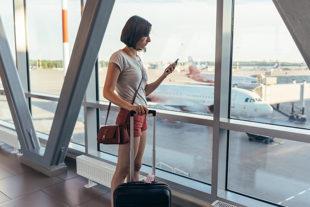 Girl standing near window at airport holding mobile phone