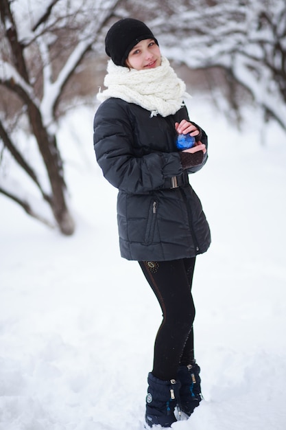 The girl stands against the background of snow-covered branches and holds in her hands a blue fur-tree toy