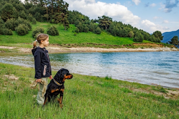 Girl stands near dog of Rottweiler breed in meadow next to lake against hilly valley with spruce forests