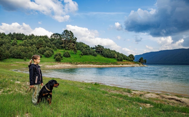 Girl stands near dog of Rottweiler breed in meadow next to lake against hilly valley with spruce forests