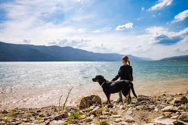 Girl stands near dog of Rottweiler breed on shore near lake against backdrop of mountain range covered with forest