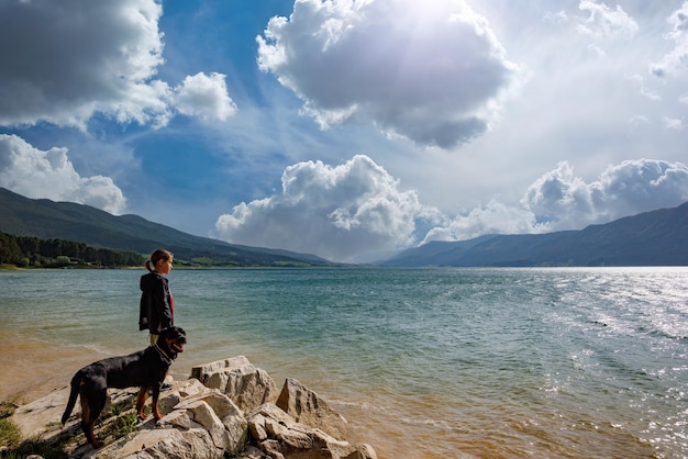 Girl stands near dog of Rottweiler breed on shore near lake against backdrop of mountain range covered with forest