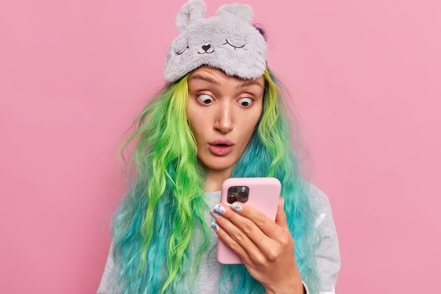  girl stares at smartphone display reads surprising news received in mesage has trendy colored hair dressed in nightwear sleepmask on forehead