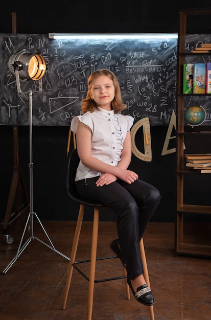 A girl in strict clothes sits on a chair near the school blackboard