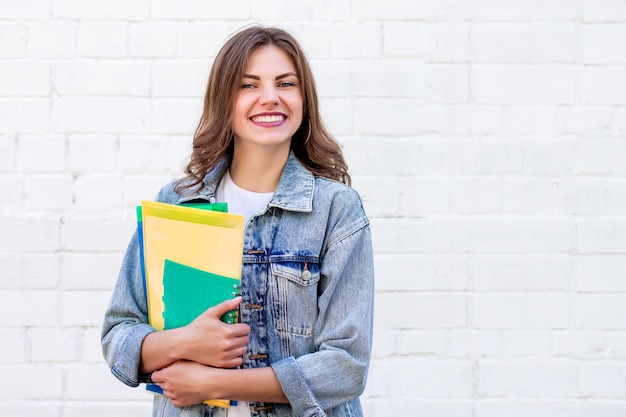 Girl student holds folders and a notebook in her hands and smiles on a background of a white brick wall, copy space