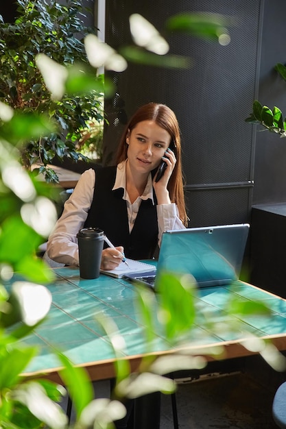 Girl student sitting at cafe table studying with laptop calling by smartphone