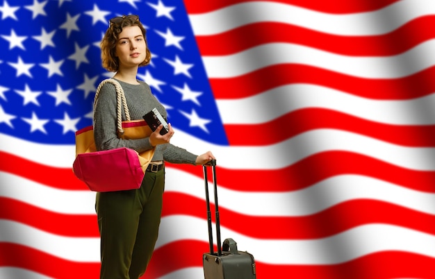 Girl student smiling, holding backpack and USA flag isolated on grey background, copy space, student exchange concept. Portrait of a cute student girl on a dark background with the flag of America.