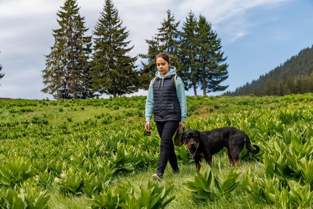 Girl in suit walks with dog of Rottweiler breed along meadow with mountain vegetation against background of trees