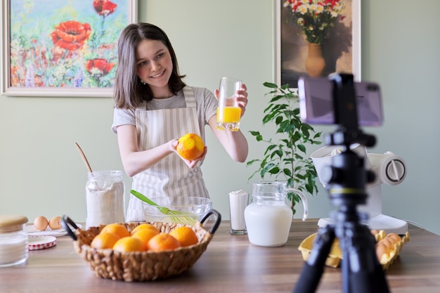 Girl teenager food blogger recording recipe for cooking orange pancakes. Woman at home in kitchen, squeezes juice, smiles and looks at camera, hobby and leisure, healthy tasty homemade food concept