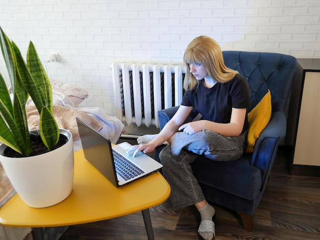 Girl teenager sits in chair near wall behind yellow table and does something in laptop