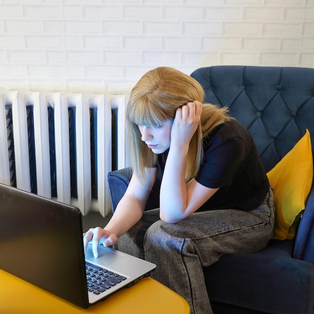 Girl teenager sits in chair near wall behind yellow table and does something in laptop