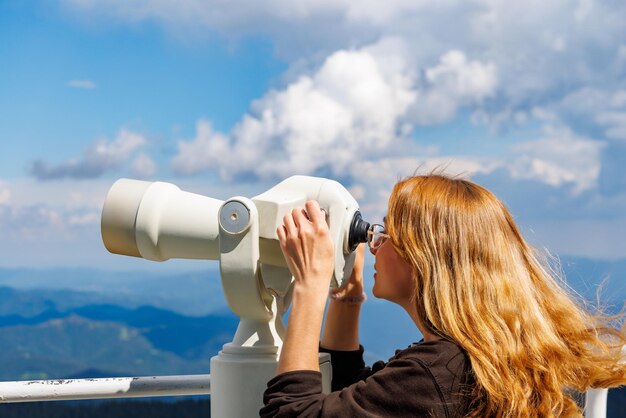 Girl tourist looks at landscapes of Rhodope Mountains and cloudy sky through telescope on high tower on top of Snezhan