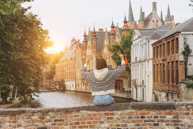 Girl tourist sits on the bridge overlooking the canal and admires the view of the picturesque and historical city of Bruges at sunset Belgium