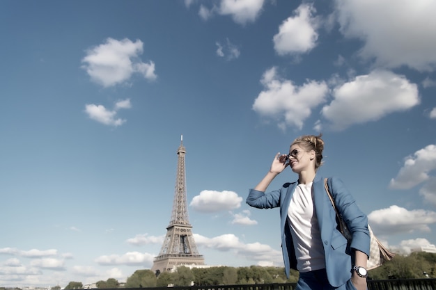 Girl traveler smile at eiffel tower in paris, france. Woman in sunglasses, jacket on sunny blue sky. Architecture, attraction, landmark. Vacation, travelling, trip. Fashion, style concept