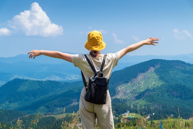 A girl traveler with a backpack stands with her arms outstretched in the mountains in summer