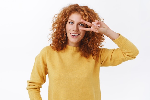 Girl trying stay positive any situation Attractive happy redhead curly woman in yellow sweater showing peace victory sign near eyes and smiling standing joyful over white background