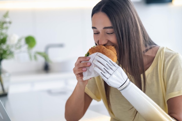 Girl using prosthetic arm while eating at home woman with disability holds croissant in artificial prosthetic limb bionic hand