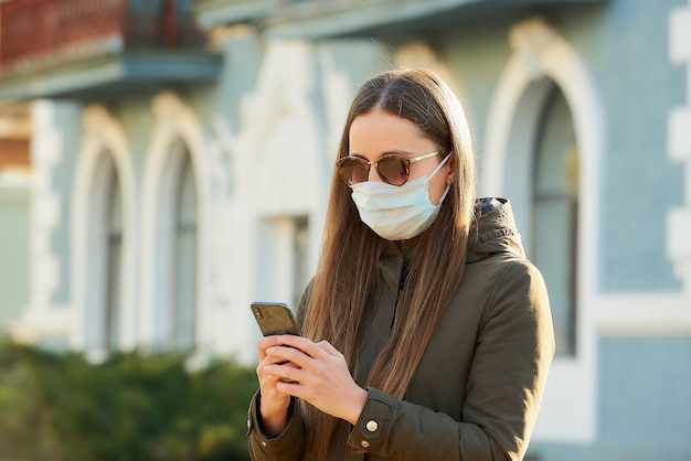 Girl using a smartphone wears a medical face mask to avoid the spread coronavirus on a city street