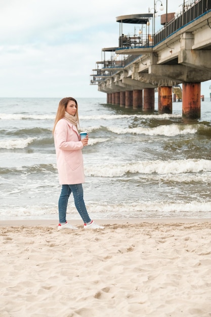 Girl walking along the seashore brunette drinks coffee girl in a pink coat sea