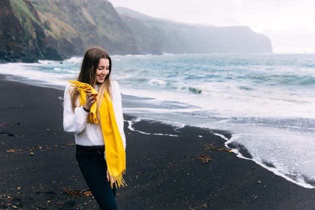 A girl walks along a volcanic sandy beach in Iceland She is wearing black pants and a white sweater with a yellow scarf