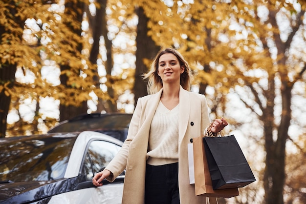 Girl walks near the car with shopping bags in hands. Modern brand new automobile in the forest.