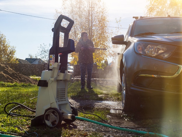 The girl washes the car at home near the garage using a pressure washer