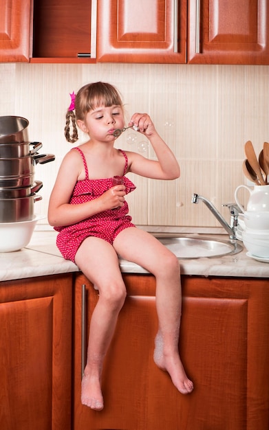 Girl washes dishes and blows soap bubbles The concept of mother's helper child development family A little girl washes white dishes at home in the kitchen Child blowing soap bubbles in the house