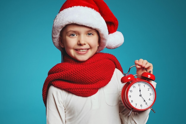 Girl wearing christmas hat and red scarf holding alarm clock isolated over