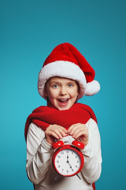 Girl wearing christmas hat and red scarf holding alarm clock isolated over