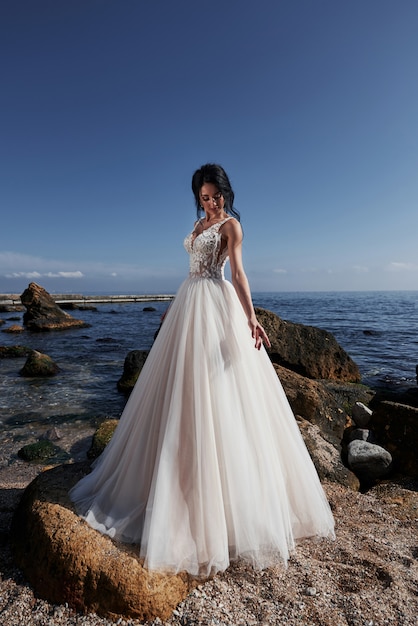 Girl in wedding luxury dress posing on sea shore. Bride on a rocks.