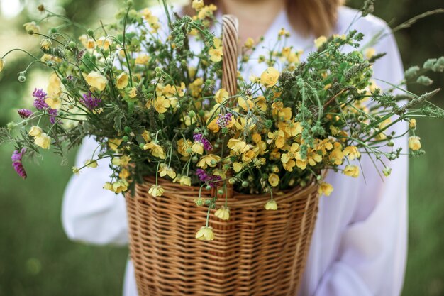 A girl in a white blouse holds a wicker basket with a bouquet of wild flowers. Summer walk in the field. midsection.