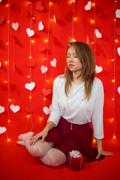 A girl in a white blouse and skirt sits on a red background with hearts. Valentine's day concept