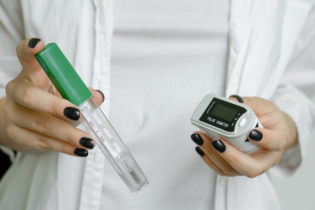 A girl in a white coat holds a thermometer to measure body temperature and a pulse oximeter covid