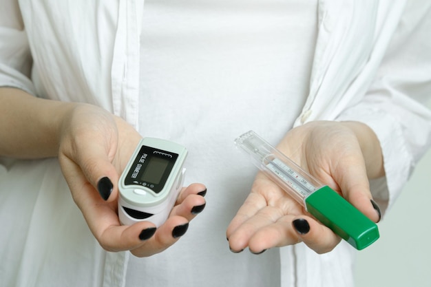 A girl in a white coat holds a thermometer to measure body temperature and a pulse oximeter covid