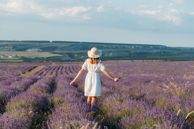 A girl in a white dress and a hat with bouquets in her hands stands back in a lavender field