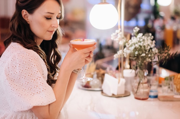 A girl in a white dress is sitting at the bar in a cafe and drinking a cocktail