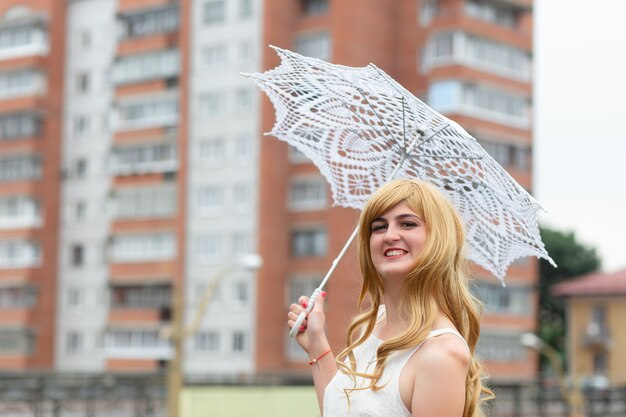 A girl in a white dress with white sun umbrella posing on city background.