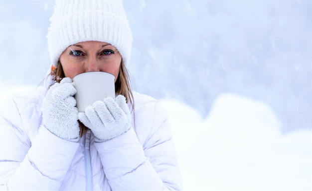 A girl in a white jacket and a white hat is holding a cup of tea in her hands during a snowfall against the background of beautiful snowdrifts