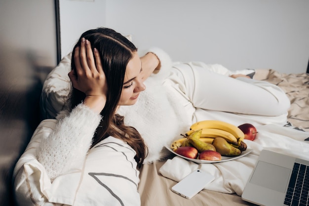 A girl in white pajamas lies in bed, resting, watching an interesting movie on her laptop and eating fruit