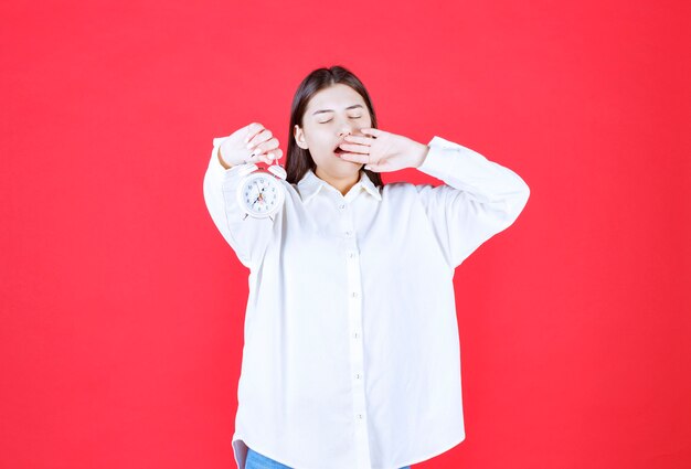 Girl in white shirt holding an alarm clock and looks sleepy