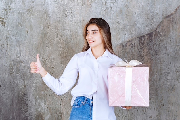 Girl in white shirt holding a pink gift box wrapped with white ribbon and showing positive hand sign