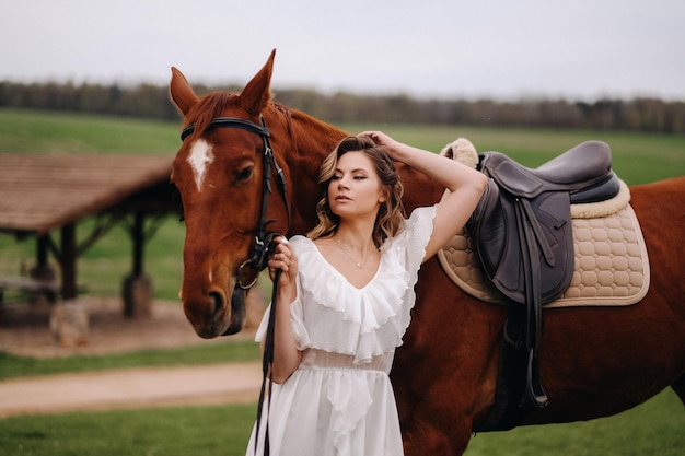 A girl in a white sundress stands next to a brown horse in a field in summer