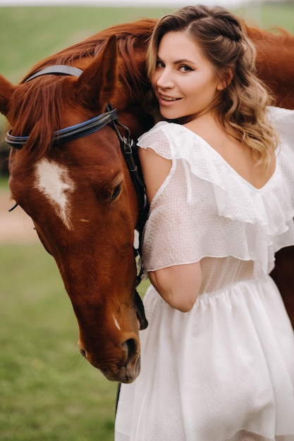 A girl in a white sundress stands next to a brown horse in a field in summer