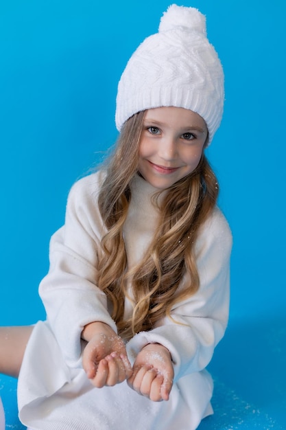 girl in a white sweater and hat blows artificial snow from the palm in the studio on blue background