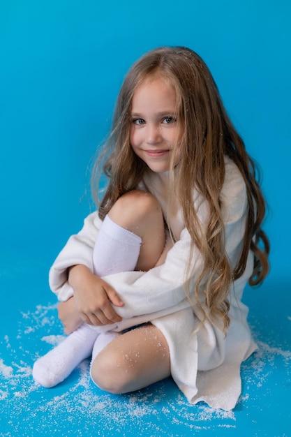 girl in a white sweater and hat blows artificial snow from the palm in the studio on blue background