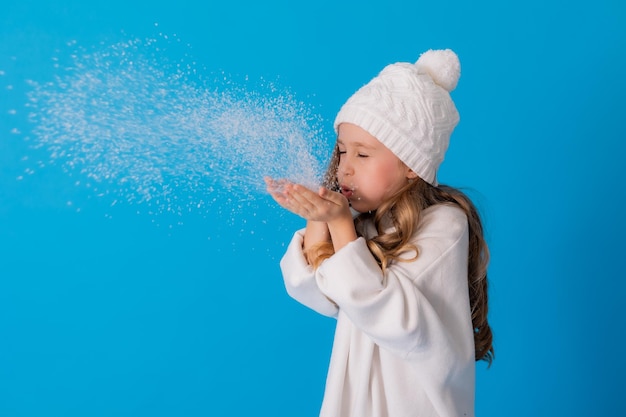 girl in a white sweater and hat blows artificial snow from the palm in the studio on blue background