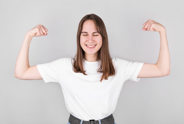 Girl in a white t-shirt with raised arms shows strong biceps, against gray wall. Sport and fitness concept