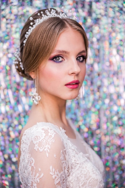 A girl in a white wedding dress with an open back makeup a hairstyle in the form of a bunch poses on glitter curtain silver background