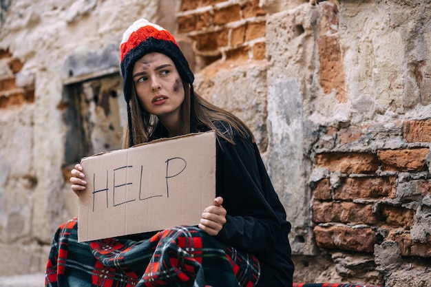 A girl who has lost her home sits dirty in the street in a blanket with a cardboard inscription Ask for help